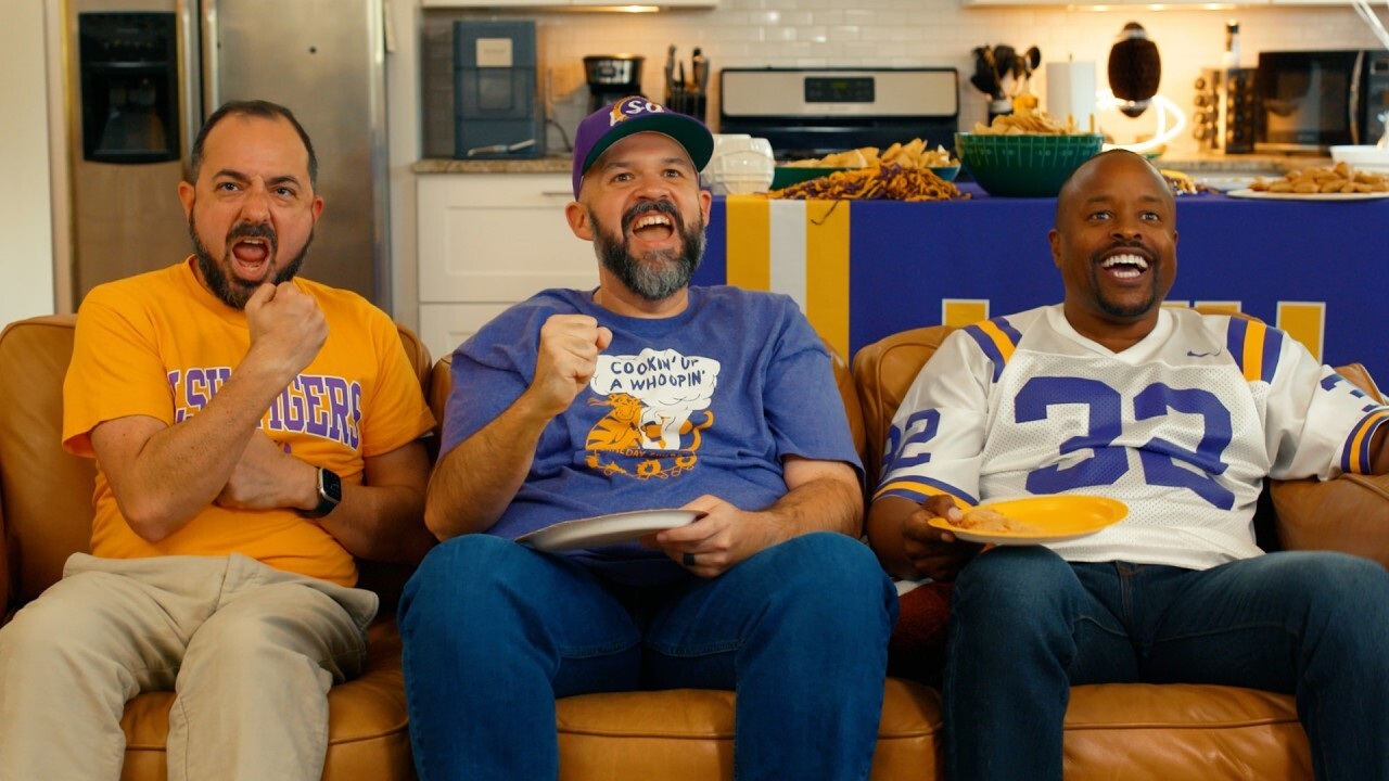 photo of three people in LSU jerseys and gear watching the TV on a sofa with snacks